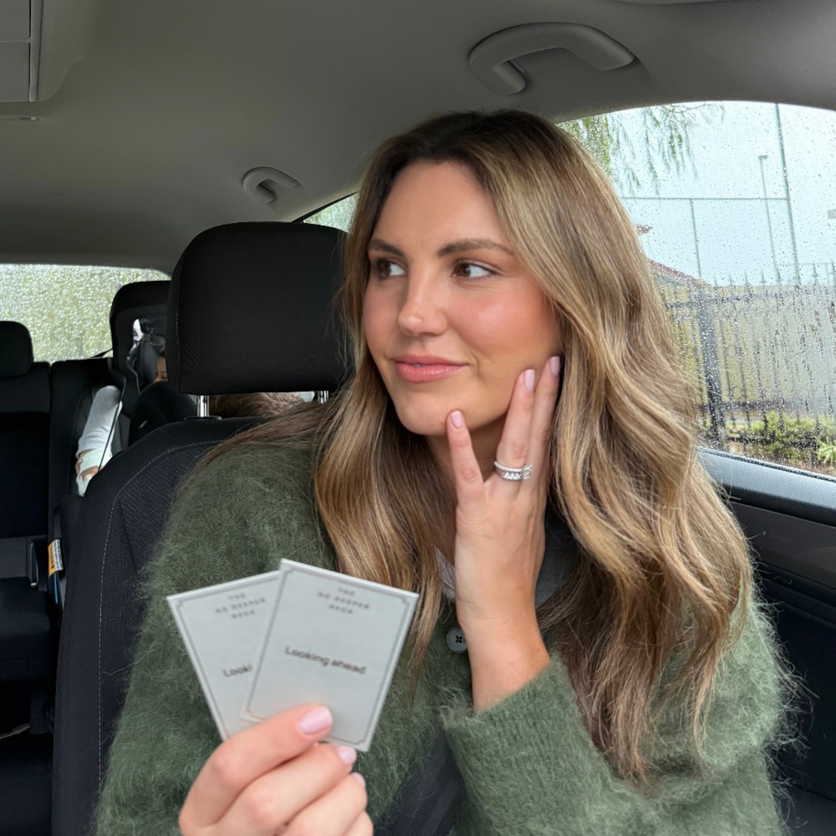 A woman in a car passenger seat holding two Go Deeper cards, smiling as she looks to the side