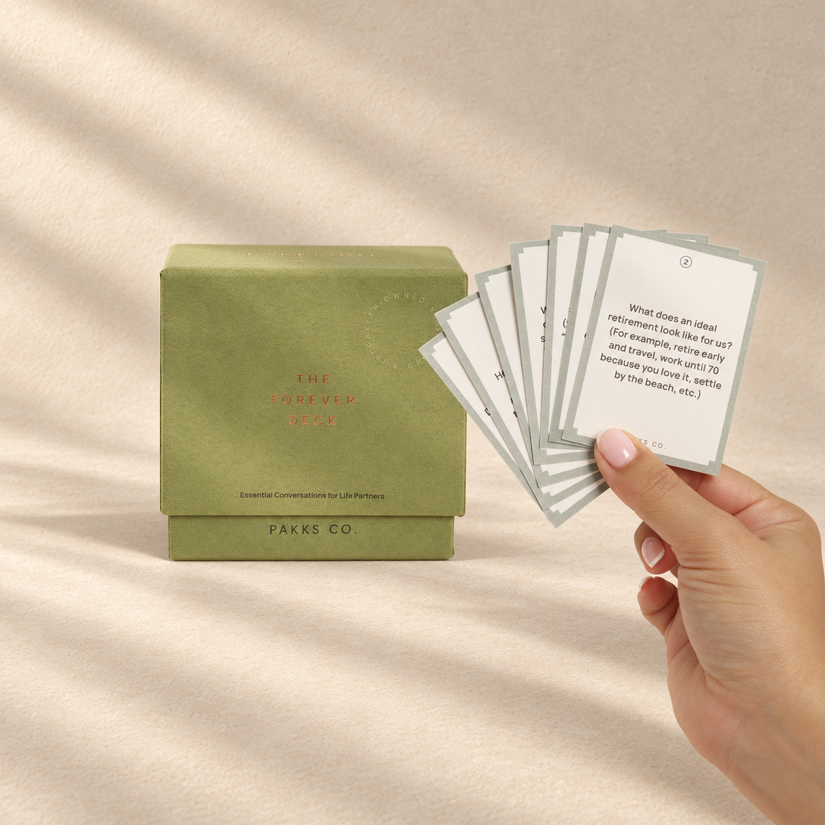 The olive-sage Forever Deck box beside a hand fanning its sage-bordered question cards in soft window light