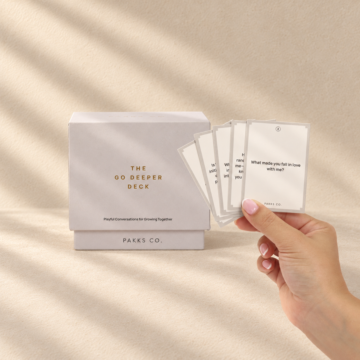 The pale grey-lilac Go Deeper Deck box beside a hand fanning its question cards in soft window light