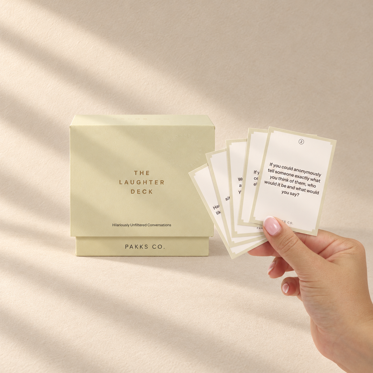 The buttercream Laughter Deck box beside a hand fanning its tan-bordered question cards in soft window light