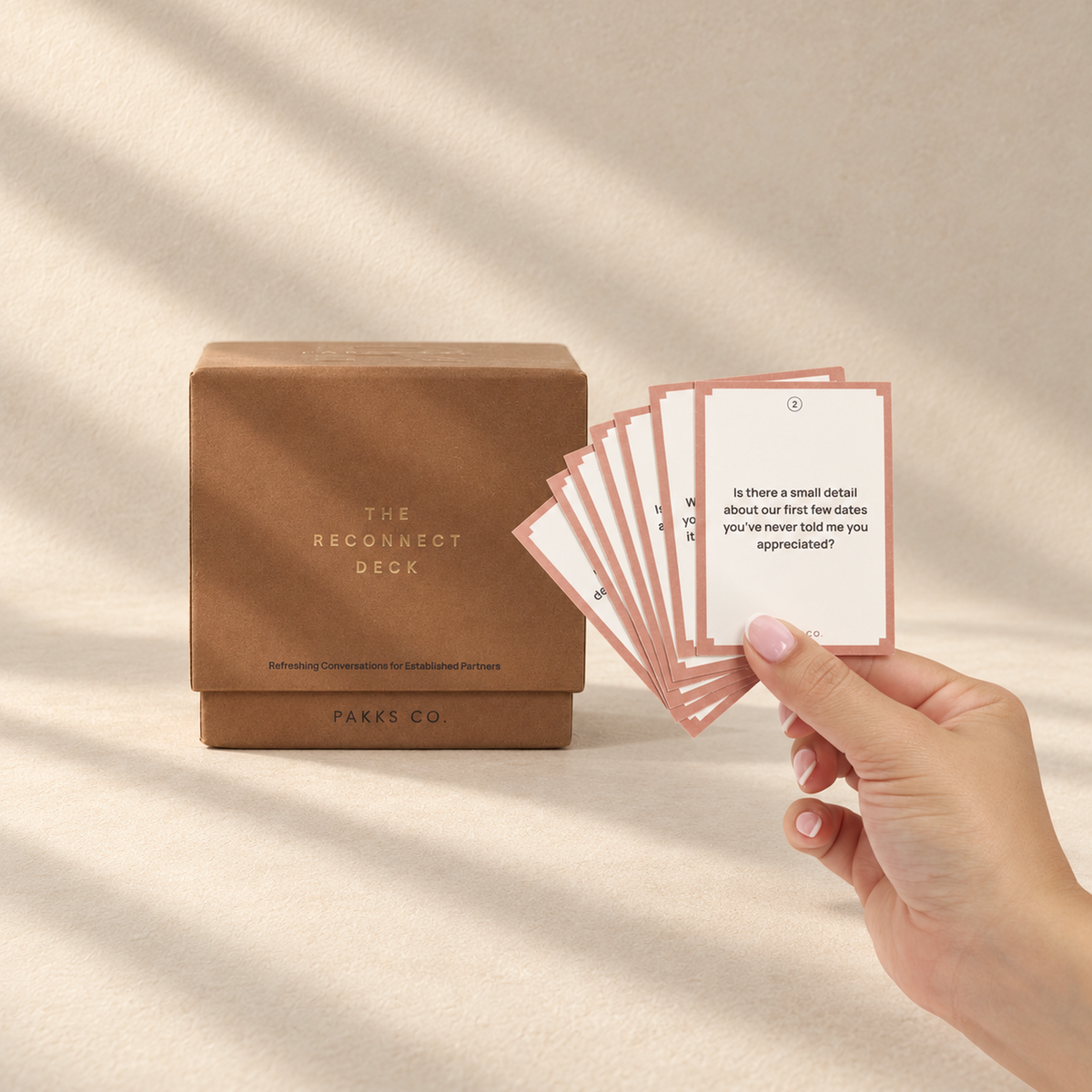 The terracotta Reconnect Deck box beside a hand fanning its pink-bordered question cards in soft window light