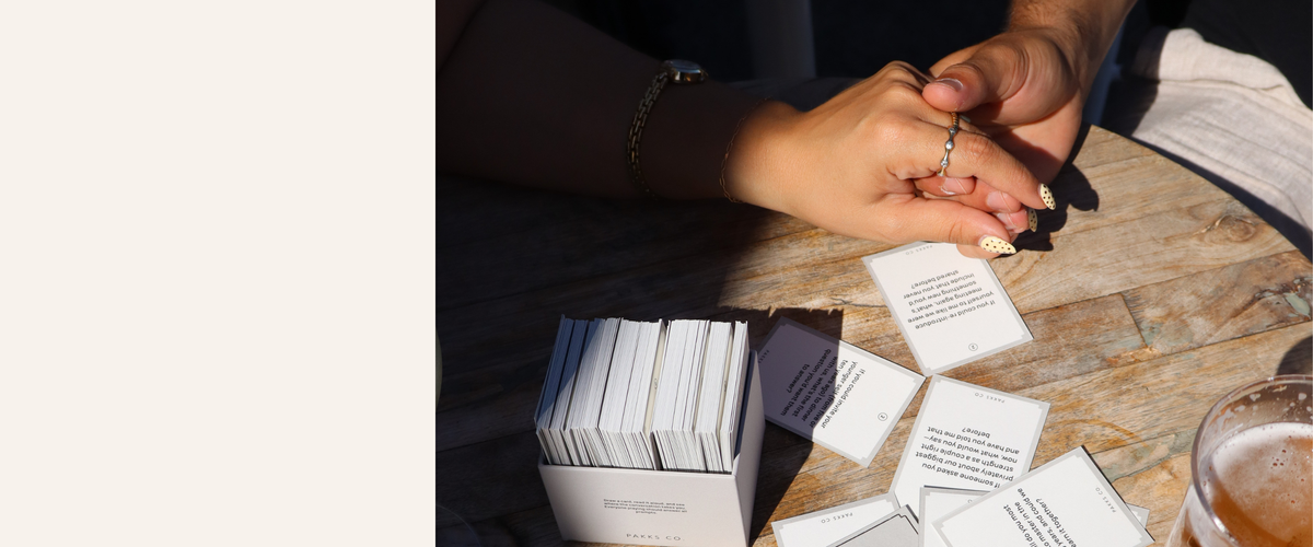 A couple's hands touching on a wooden table beside scattered Pakks cards, an open card box and a beer