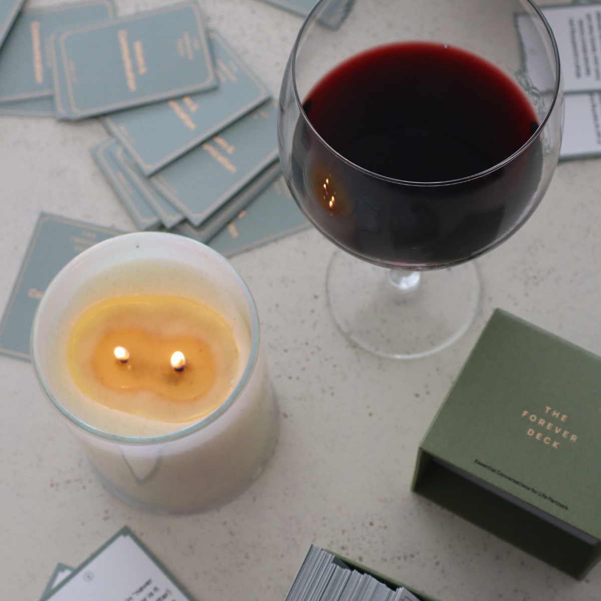A lit candle, a glass of red wine and the Forever Deck box with its cards scattered on a table