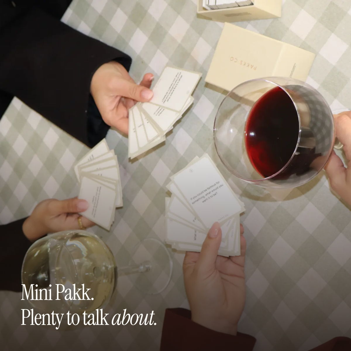 Friends passing Laughter Deck cards over a gingham table with wine, captioned "Mini Pakk. Plenty to talk about."