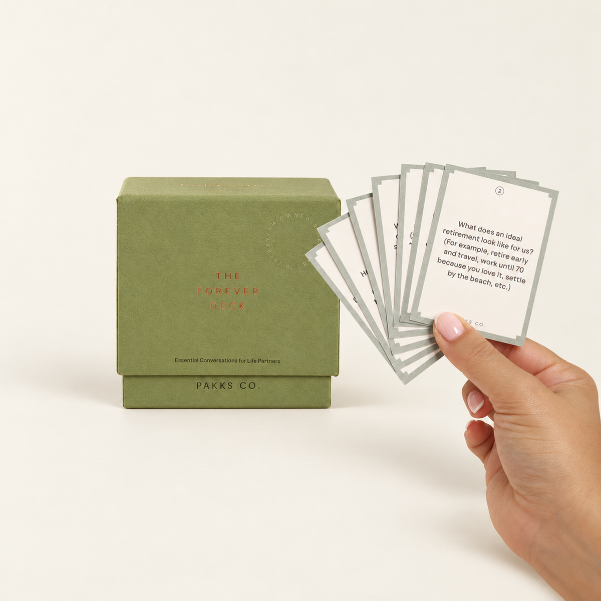 The Forever Deck: an olive-sage box beside a hand fanning sage-bordered question cards on an ivory background