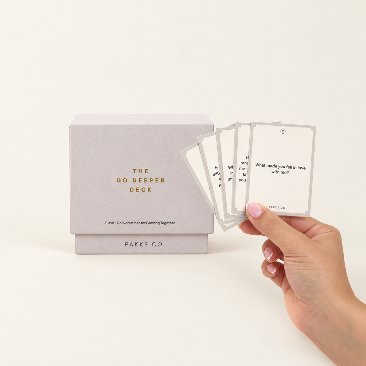 The pale grey-lilac Go Deeper Deck box beside a hand fanning cards, front card "What made you fall in love with me?"