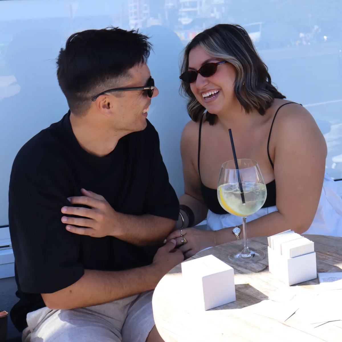 A couple in sunglasses laughing at an outdoor table with a drink and white card boxes