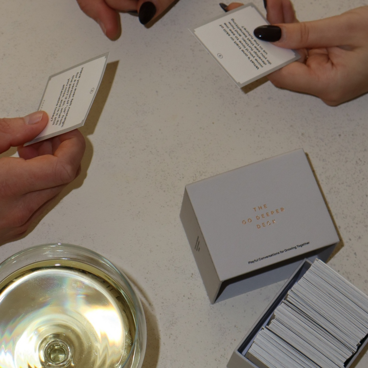 Overhead view of hands holding Go Deeper cards around the open box and a glass of white wine