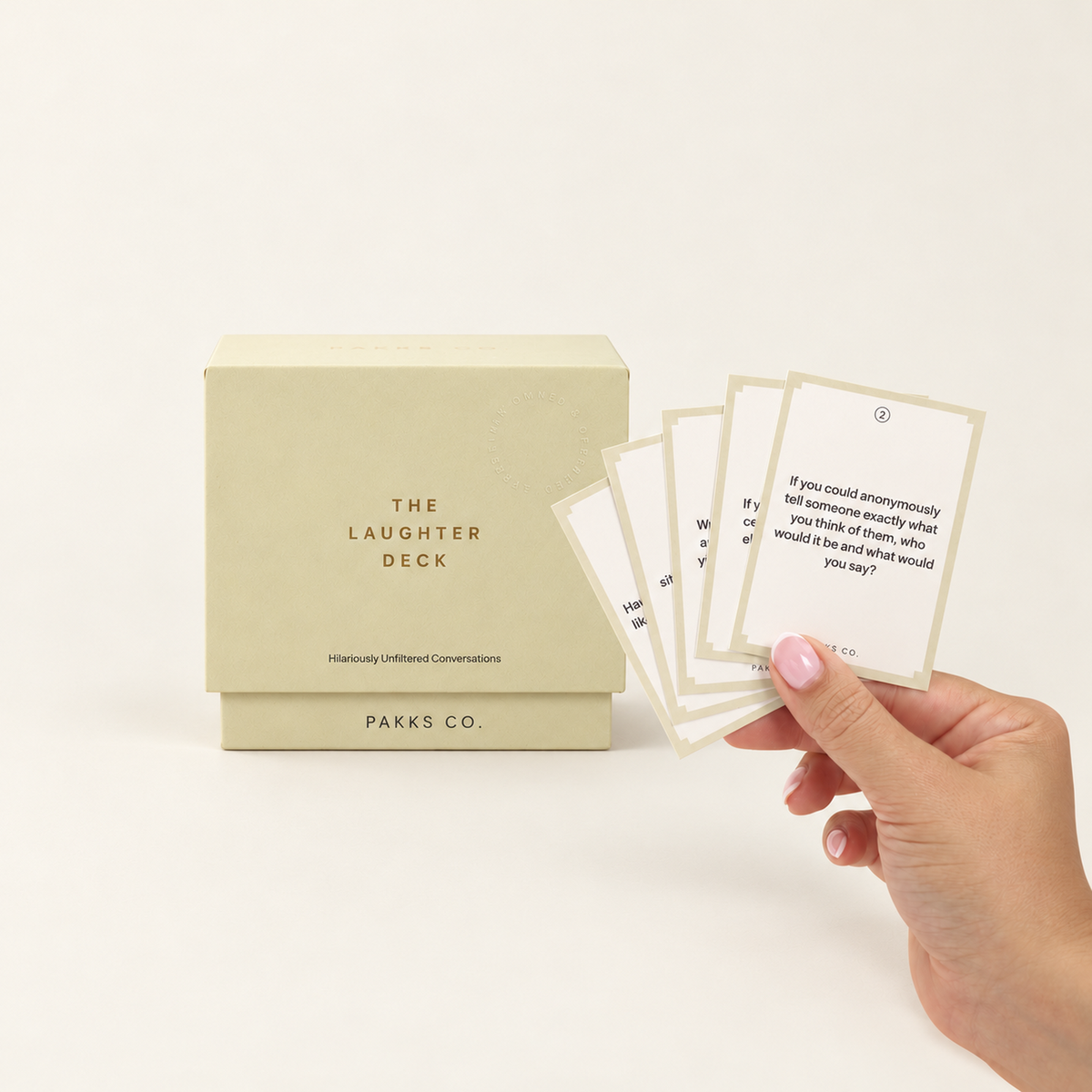 The Laughter Deck: a buttercream box beside a hand fanning tan-bordered question cards on an ivory background