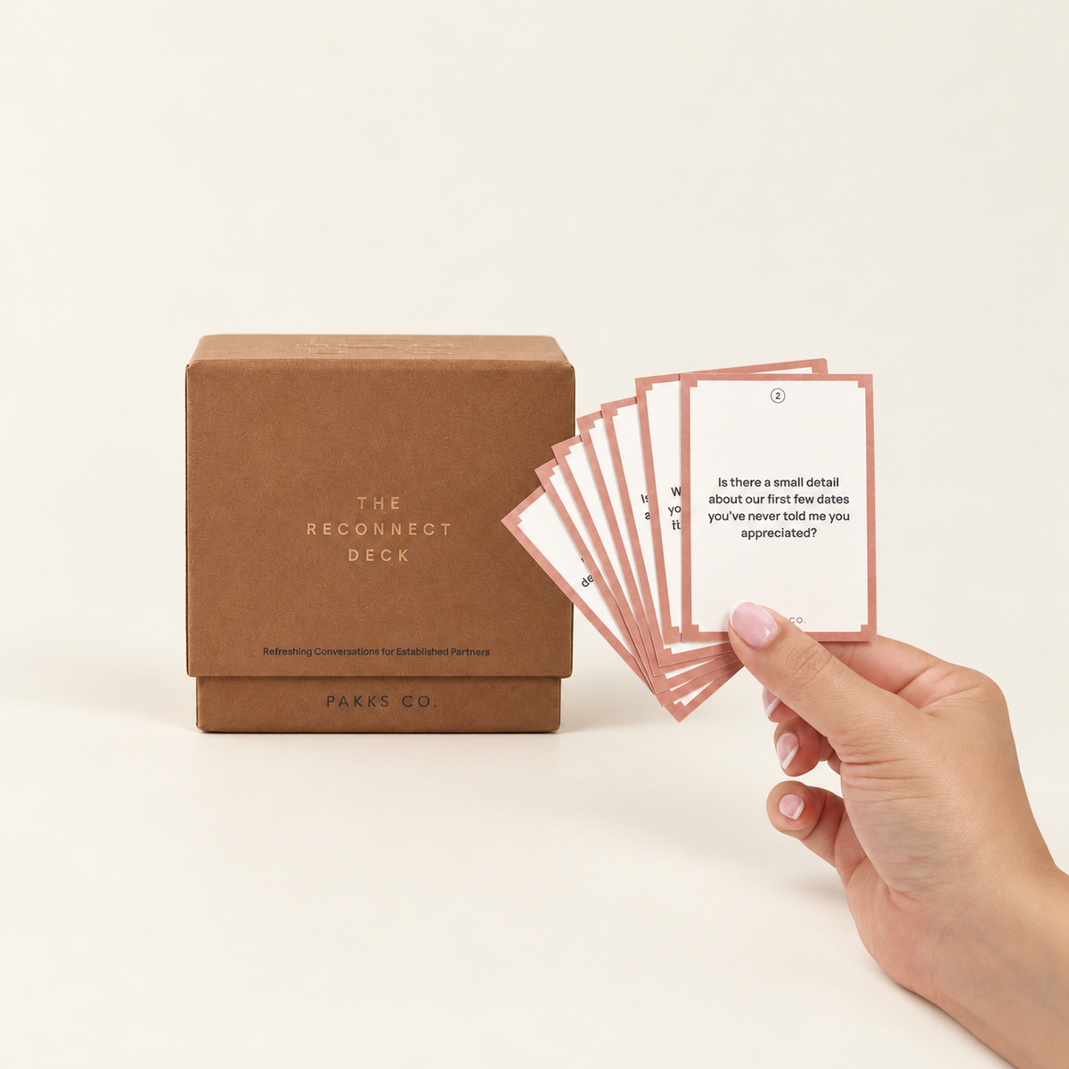 The Reconnect Deck: a terracotta box beside a hand fanning pink-bordered question cards on an ivory background