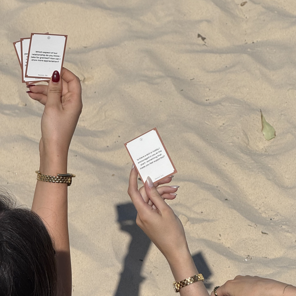Two hands holding up Reconnect question cards against sunlit beach sand