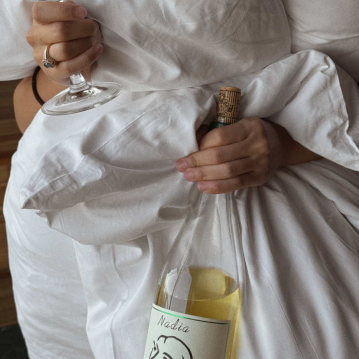 Someone wrapped in white bedsheets holding a wine glass and a bottle of white wine